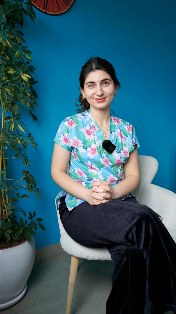 Healthcare professional in floral scrubs seated indoors with a plant background.